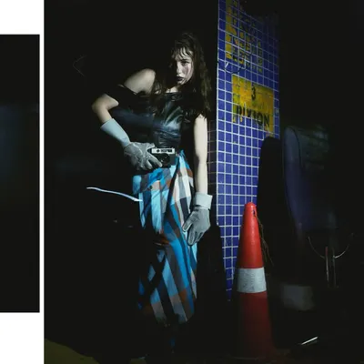 A full-length shot from the 'Peek-a-boo' series, set in a gritty, industrial-looking location. The harsh lighting and dark shadows create a mysterious, noir atmosphere.
