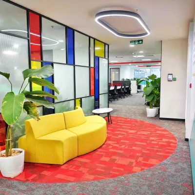 A vibrant lounge area in the Mindsprint office, featuring a curved yellow sofa set against a Mondrian-inspired colorful glass partition. The circular red carpet defines the space, making it a perfect spot for informal chats and breaks.