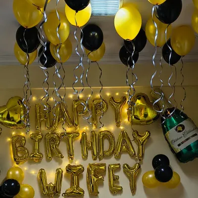 A classic birthday surprise for a loved one. This room decoration features gold and black balloons on the ceiling, "Happy Birthday Wifey" foil letters, and champagne bottle foils for a celebratory touch.