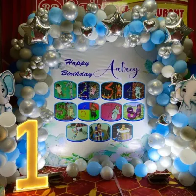 An adorable elephant-themed first birthday party backdrop. The setup features a balloon arch in blue, silver, and white, a large light-up number '1', and a personalized backdrop with the baby's monthly milestone photos.