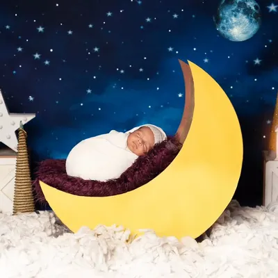 A beautiful portrait of a newborn swaddled in white, sleeping on a large yellow moon. The deep blue, star-filled background and miniature set pieces complete this enchanting celestial scene.
