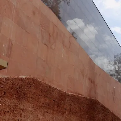 A detail where the raw, earthy texture of the red sandstone wall meets a sleek, reflective glass facade. This juxtaposition of materials is a core element of the design, celebrating both natural texture and modern precision.