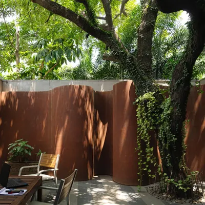 An outdoor seating area at the KEUS experiential hub, nestled against a curving Corten steel wall and shaded by a mature tree. This space is designed as a breakout zone for work or relaxation, blending industrial materials with natural elements.