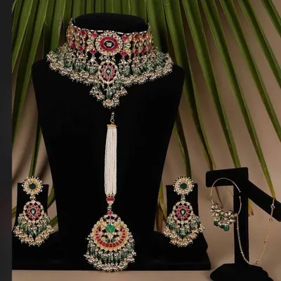 A complete bridal jewellery set available for rent. This includes a heavy choker, matching earrings, a maang tikka, and a nath, all with beautiful red and green stones.