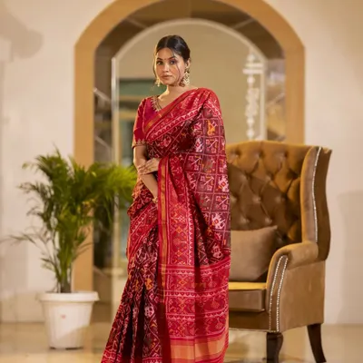A full-length shot of a model in a traditional red Patola saree. The elegant standing pose in a well-appointed room highlights the drape and richness of the fabric, perfect for festive wear collections.