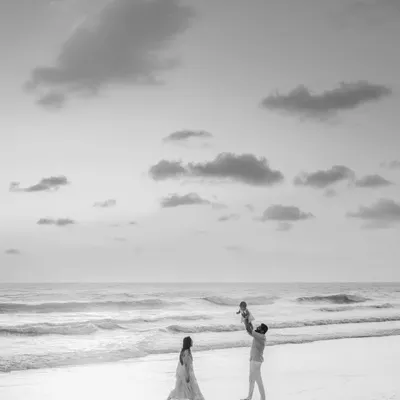 A striking black and white photo of a family on the beach. The father joyfully lifts their child into the air while the mother walks nearby, creating a dynamic and artistic family portrait.