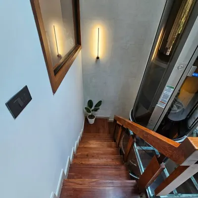 A top-down view of the stairwell, showcasing the sleek, all-glass vacuum elevator alongside Indonesian walnut stair treads. Ambient wall lighting and a touch of greenery complete this statement of luxury and smart living.