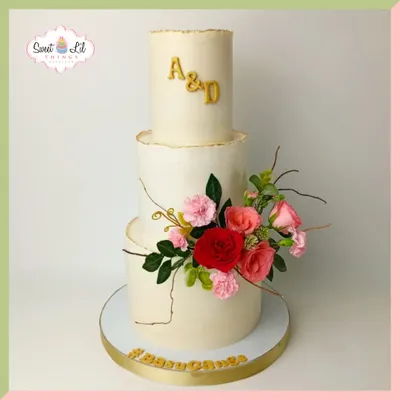 A timeless and elegant three-tiered wedding cake. The clean, white tiers are personalized with the couple's initials and adorned with a beautiful, fresh floral arrangement in shades of pink and red.