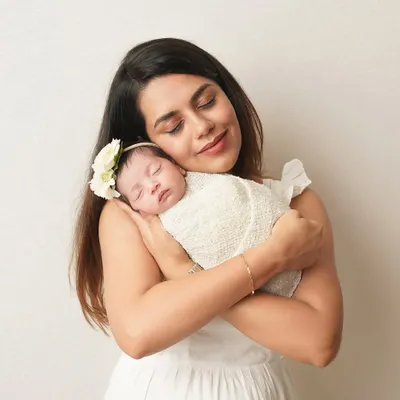 A mother in a white dress gently holds her sleeping newborn, who is swaddled in a textured white wrap with a delicate floral headband. The all-white theme creates a pure and serene portrait.