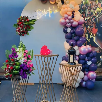 A stylish "Bride to be" setup with a modern, edgy vibe. The decor features a white backdrop with a neon sign, an asymmetrical balloon arch in rich tones, and fresh floral arrangements on gold stands.