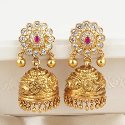 Classic gold-toned jhumkas featuring a floral stud set with white stones and a central ruby-colored gem. The bell is intricately carved, offering a timeless and versatile design.