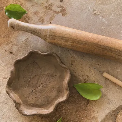 A hand-built bowl with a leaf impression, sitting next to a wooden rolling pin. In its raw, unfired state, you can see the delicate details and the soft texture of the natural clay.