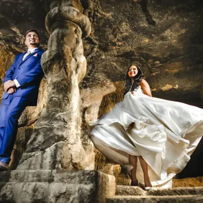 A dynamic post-wedding portrait taken inside ancient rock-cut caves. The natural light and rugged texture of the stone create a dramatic backdrop, highlighting the couple's joy and the bride's flowing dress.