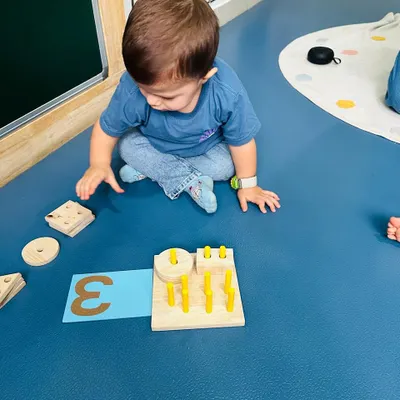 A toddler explores shapes and numbers with a wooden puzzle. My training emphasizes using natural, tactile materials to introduce foundational concepts like numeracy and geometry.