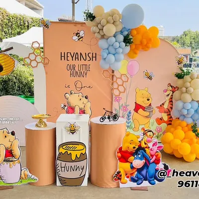 A delightful Winnie the Pooh "Our Little Hunny" first birthday party. This outdoor setup features character cutouts, honey-pot-themed plinths, and a large number '1' filled with balloons in a pastel color scheme.