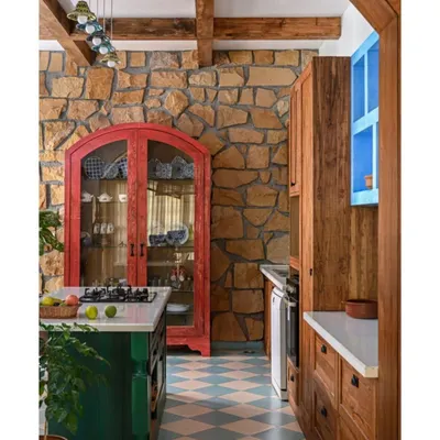 This kitchen is designed to feel like it's from another time, miles away from fleeting trends. We used a bold color palette with a green island, a red crockery cabinet, and a checkered floor, all set against a rugged stone wall and exposed wooden beams. Every element is chosen to create a warm, rustic, and deeply personal cooking space.