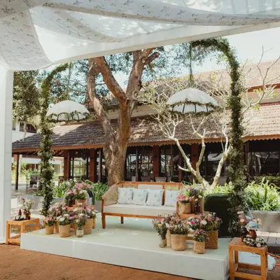 A minimalist Mehendi stage designed for a laid-back Goan wedding. A simple wooden bench is framed by two delicate floral arches and whimsical hanging umbrellas, creating a look that is both modern and dreamy.