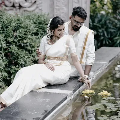 A serene and romantic pre-wedding photo of a couple in traditional white South Indian attire, sitting by a lily pond, capturing a moment of quiet intimacy.
