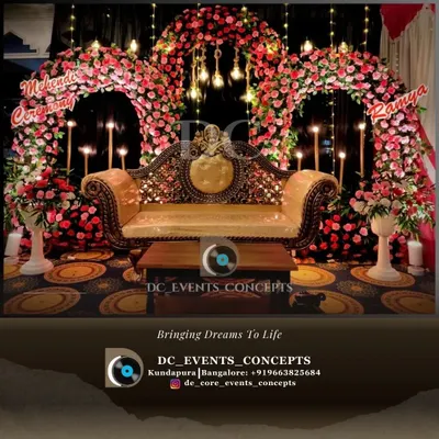 An extravagant Mehendi stage with twin floral arches in vibrant pink and orange. The traditional gold sofa and hanging lights complete this lavish setup, perfect for a grand pre-wedding celebration.