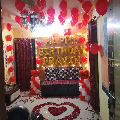 A complete romantic birthday surprise package. This room decoration includes red and white balloons, fairy lights, 'Happy Birthday' foil letters, and a heart-shaped rose petal arrangement on the floor.