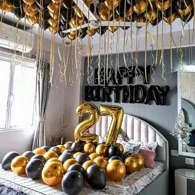 A stylish 27th birthday room decoration with black and gold balloons covering the bed and ceiling. The setup includes large number '27' foil balloons and 'Happy Birthday' letters on the wall.