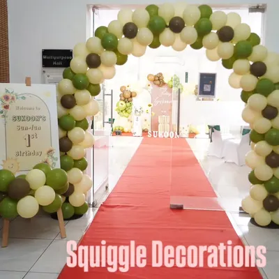 An inviting entrance for a sunflower-themed first birthday party. The decor includes a balloon arch in rustic colors, a red carpet, and a personalized welcome sign.