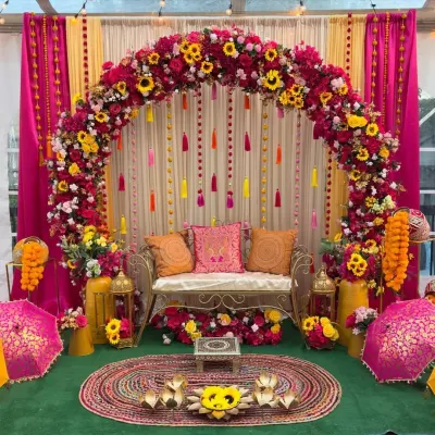 A full view of our popular Haldi and Mehndi stage decor. The setup features a dense floral arch, a comfortable bench with decorative cushions for the bride, and traditional elements like decorated umbrellas and brass lanterns.