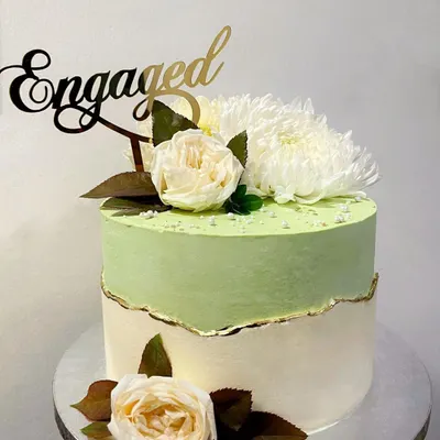 This fault-line cake was designed for an engagement party. It features a soft pistachio green top section over an off-white base, with the "break" highlighted by a delicate gold leaf edge. The design is completed with an "Engaged" topper and fresh white flowers.