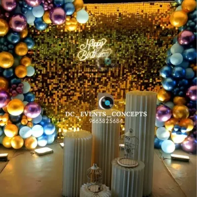 A glamorous birthday photo booth featuring a gold shimmer wall framed by a colorful balloon arch. The setup is completed with decorative pedestals and a custom neon "Happy Birthday" sign.