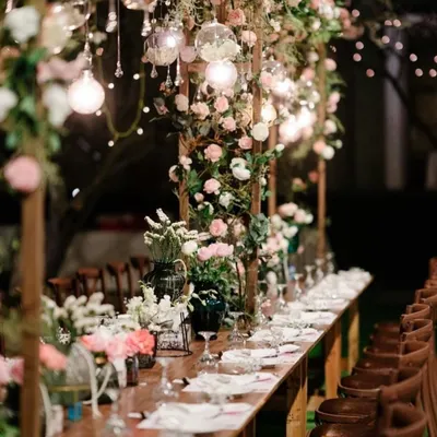 An enchanting long table setup for an evening reception, with a wooden structure overhead covered in flowers and hanging lights.
