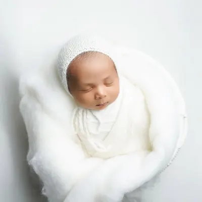 An 18-day-old baby boy, swaddled in soft white wool and a delicate bonnet. This portrait highlights the serene and pure essence of a newborn, captured with a minimalist approach.