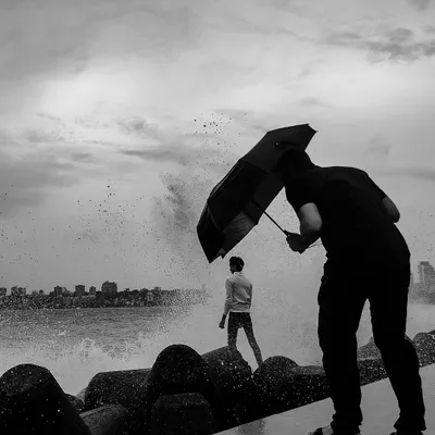 A dramatic black and white shot of a man holding an umbrella against the wind at Marine Drive, as waves crash against the tetrapods. The monochrome treatment enhances the raw power of the sea and the drama of the moment.