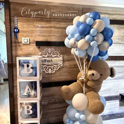 This welcome home arrangement for a baby boy includes a teddy bear holding a bouquet of blue and beige balloons, positioned next to transparent 'BABY' blocks filled with smaller matching balloons.