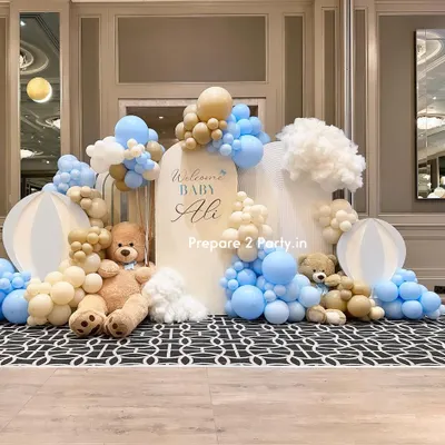 A luxurious and dreamy welcome home setup for "Baby Ali" in a grand hall. The design features soft blue, beige, and white balloons creating a cloud-like effect around arched backdrops, complete with large teddy bears and orb props.