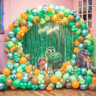 A roaring good time with a dinosaur-themed birthday! This setup features a vibrant green and orange balloon arch, a neon sign, and friendly dinosaur character cutouts.