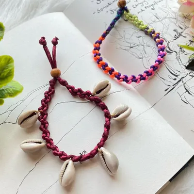 A pair of my beach-inspired anklets. One design features a deep red braided band adorned with natural cowrie shells for a classic beachy look, while the other is a vibrant, multi-colored braided anklet. Both are adjustable for a comfortable fit.