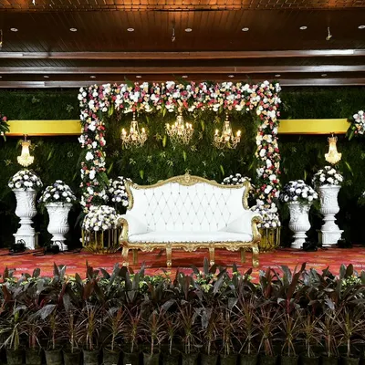 An elegant indoor reception stage featuring a classic white couch set against a lush green wall. The floral arch, Roman pillars, and warm chandeliers come together to create a look of timeless sophistication.