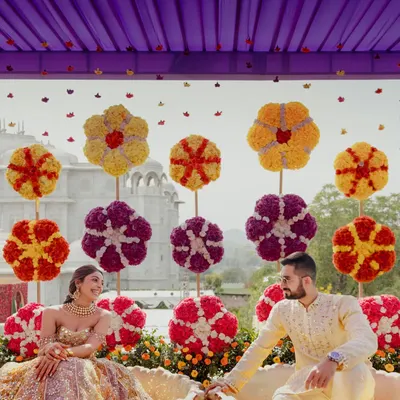 The bride and groom seated for their Haldi ceremony against a backdrop of giant, handcrafted floral lollipops. The playful design and vibrant colors under a lilac canopy create a joyful and unique setting.