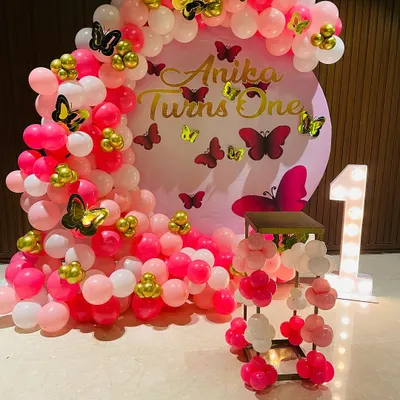 A beautiful butterfly-themed first birthday setup in shades of pink. The circular backdrop is framed by a dense balloon arch, with gold and pink butterfly stickers adding detail throughout the design.