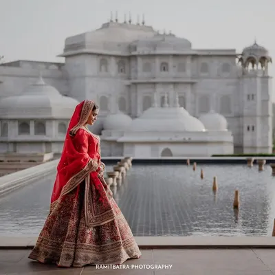 A bride in her stunning red lehenga stands against the magnificent architecture of Fairmont Jaipur, a portrait of regal grace.