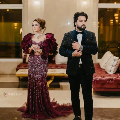 A stylish, full-length portrait of the couple at their reception, showcasing the bride's dramatic ruffled gown and the groom's classic tuxedo.