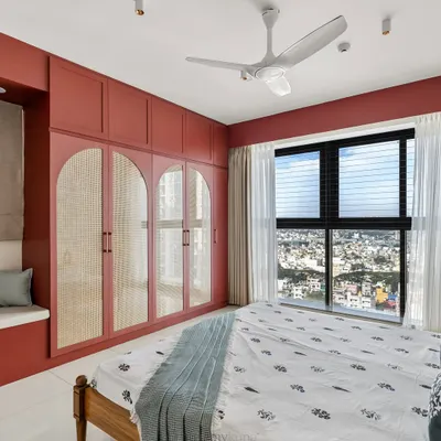 This master bedroom in a Shapoorji Pallonji flat features a bold burgundy red wardrobe with arched cane detailing. It proves that a strong color choice can create a warm, inviting, and highly functional space, complete with a cozy bay window seat.