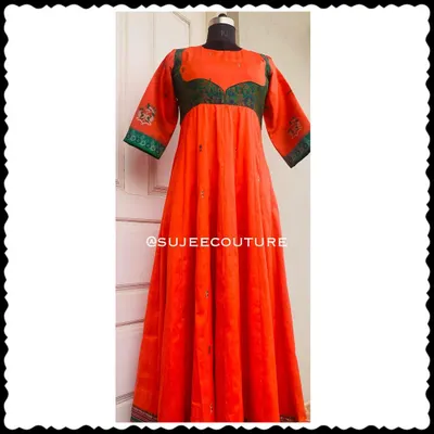 A full-length view of a custom Anarkali-style gown. This vibrant orange dress features a contrasting green yoke made from brocade fabric, with subtle embroidery on the sleeves and hem.