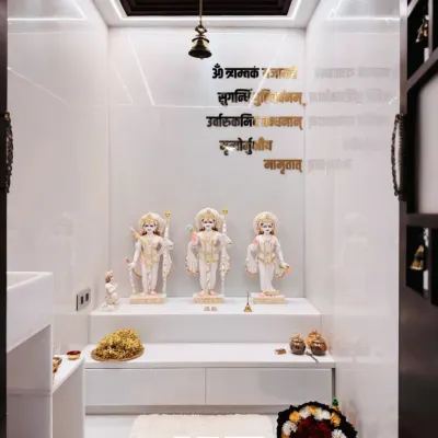 This minimalist home mandir creates a sanctuary of peace. The design features pristine white marble idols, a clean white platform with storage, and the sacred Gayatri Mantra in elegant gold lettering on the wall.