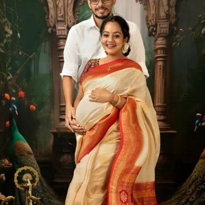 A classic couple portrait from a traditional maternity session. The mother-to-be is radiant in a cream and red silk saree, standing with her partner against a backdrop inspired by Indian art, complete with peacocks.