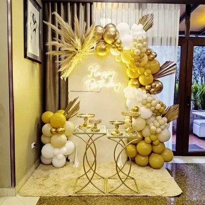 This full boho-glam setup features a half-arch balloon garland in gold and white, accented with fluffy pampas grass. The 'Happy Birthday' neon sign adds a modern touch, while the gold dessert tables provide a stylish space for the cake and treats.