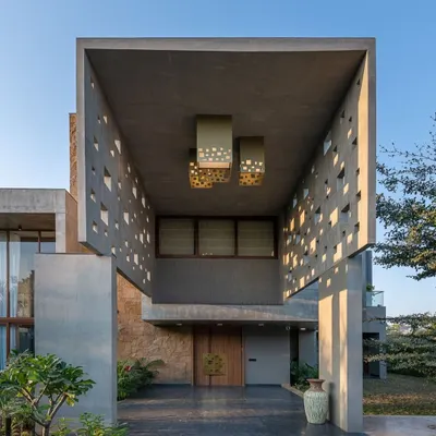 The entrance to this home is a study in Brutalist-inspired design. A massive concrete canopy with geometric cutouts creates a dramatic play of light and shadow, guiding you toward the warm wooden front door.
