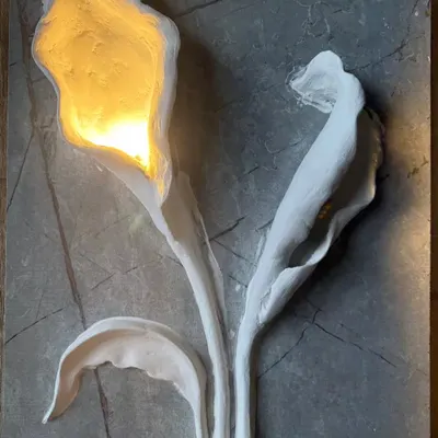 This 3D wall sculpture features calla lilies with a warm light glowing from within. Set against a grey marble-effect panel, the piece is both a work of art and an elegant light fixture.