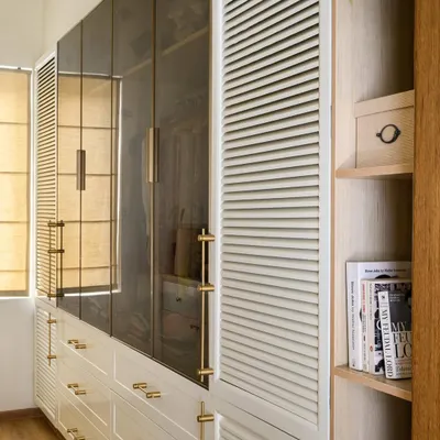 This elegant walk-in closet in our 'House of Kismish' project features a blend of white PU-finished drawers, tinted glass shutters with brass handles, and louvered panels for ventilation.