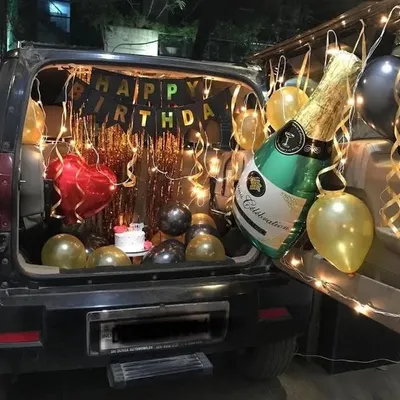 A birthday surprise in a car boot, complete with a mini cake, a foil curtain, fairy lights, and a large champagne bottle balloon.